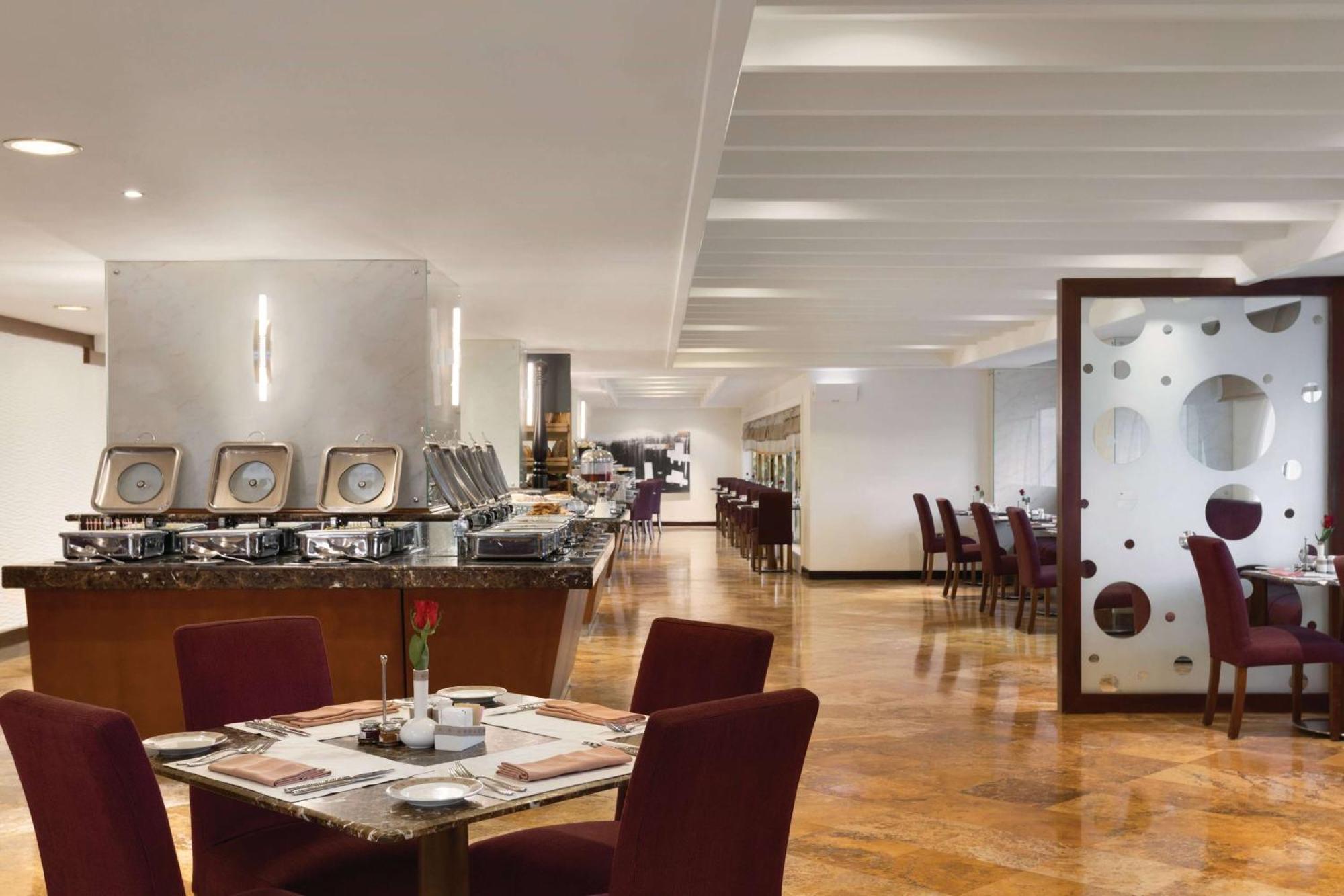 Grand Regency Doha, Trademark Collection By Wyndham Экстерьер фото The photo shows a modern restaurant interior. There are several tables with maroon chairs arranged throughout the space. One table is set with a small vase containing a flower and dining utensils. Along one side, there is a buffet area with a sleek c