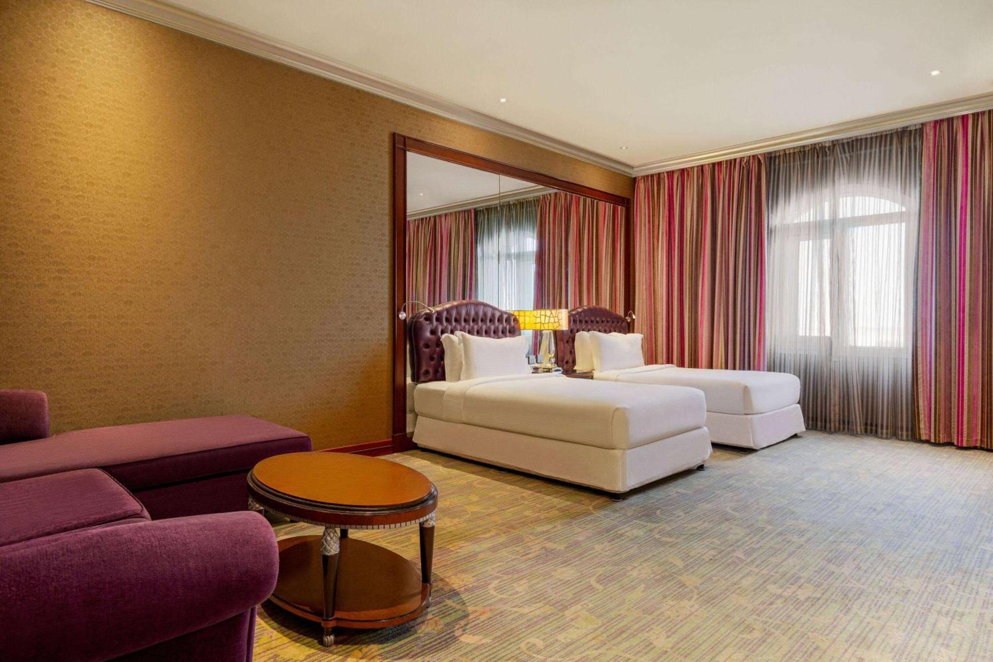 Grand Regency Doha, Trademark Collection By Wyndham Экстерьер фото The photo shows a hotel room featuring two beds with white bedding, a large mirror on one wall, and a window dressed with sheer and curtain panels. There is a small round table in the center of the room and a plush purple armchair nearby. The overall
