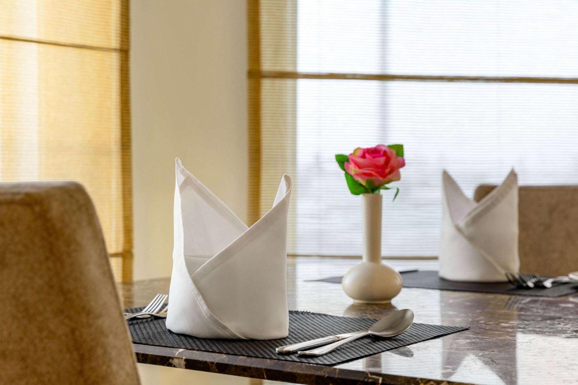 Grand Regency Doha, Trademark Collection By Wyndham Экстерьер фото The photo shows a neatly arranged dining table. On the table, there are two cloth napkins folded in a decorative way, one at each place setting. Silverware, including a knife and fork, is placed next to each napkin. In the center of the table, there 