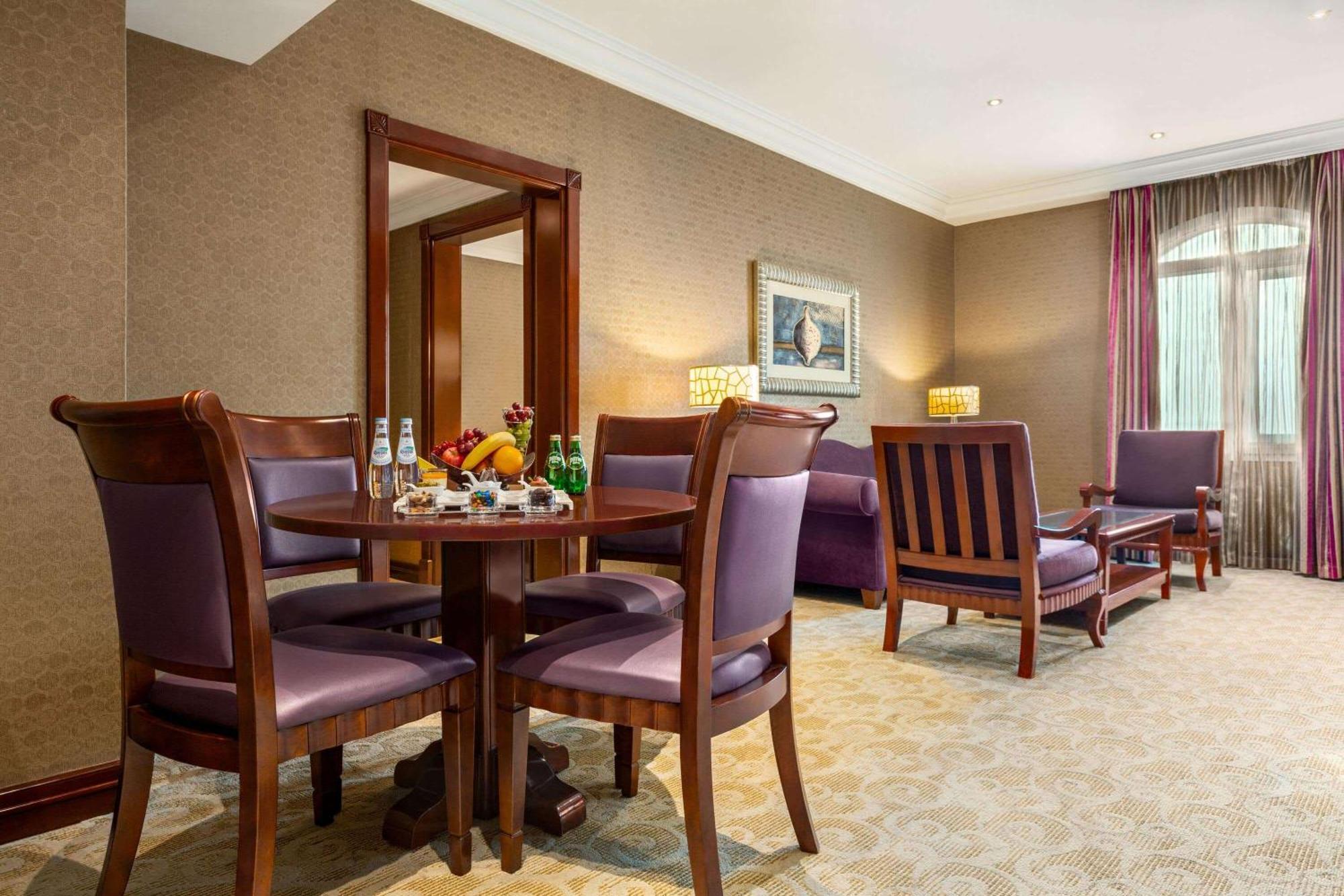Grand Regency Doha, Trademark Collection By Wyndham Экстерьер фото The photo depicts a well-furnished hotel room or lounge area. In the foreground, there is a round table surrounded by four purple upholstered chairs, with a selection of fruits and beverages on the table. To the right, there are two wooden armchairs,